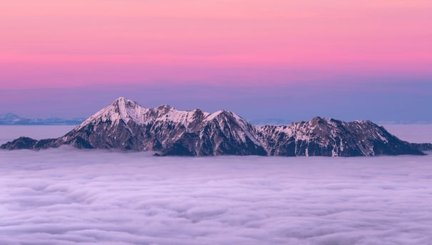 a snow covered mountain range poking above the clouds shrouded in pink purple evening sky