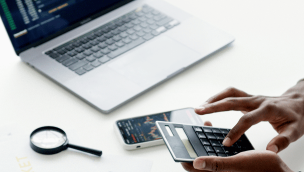 Employee conducting financial analysis using a calculator next to a laptop with charts.