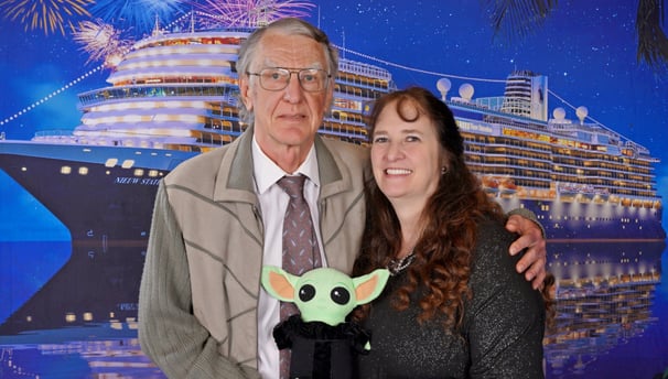 Man and Woman standing in front of a Cruise Ship holding a green stuffed animal between them.