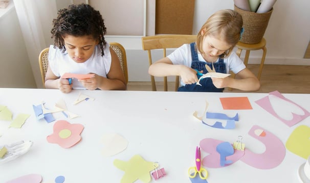 Two young girls cutting paper with scissors whilst making crafts at a table.