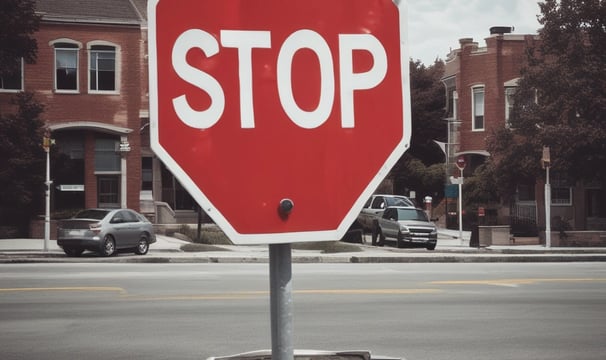 A person in an orange uniform and straw hat is cleaning a sidewalk with a broom. The walkway is lined with lush green bushes and large trees provide shade. Vehicles are parked by the roadside. Construction workers are visible further down the walkway with signage placed nearby, indicating an ongoing operation.