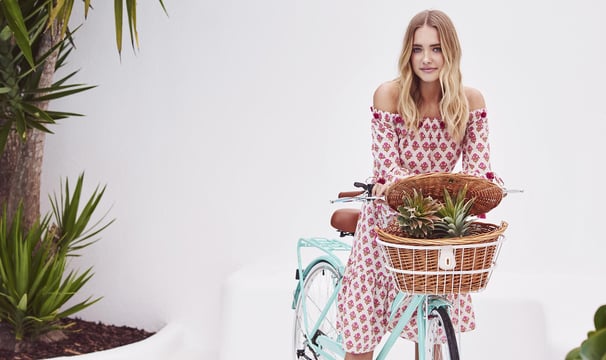 Jeune femme en robe à motifs assise sur un vélo Reid turquoise avec panier en osier contenant des pl