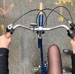 Vue du guidon d’un vélo bleu en mouvement sur une route, mains au guidon et jambes visibles.