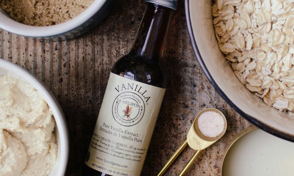 A bottle of vanilla extract beside bowls of rolled oats, brown sugar, and dough