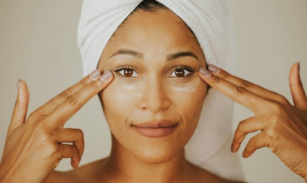 Woman with towel-covered hair applying a skincare product around her eyes