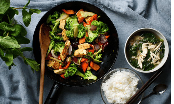 Un wok avec un mélange de légumes et un bol de riz évoquant un repas sain et équilibré asiatique.
