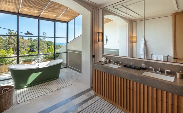 Bathroom with freestanding green tub, double vanity, and floor-to-ceiling garden view windows.