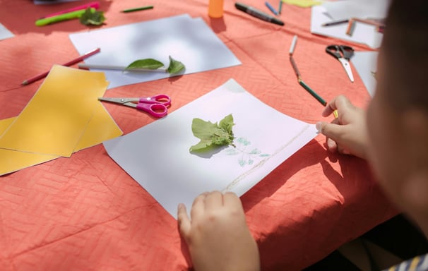 a child's hands are holding scissors and paper and leaves are on the table