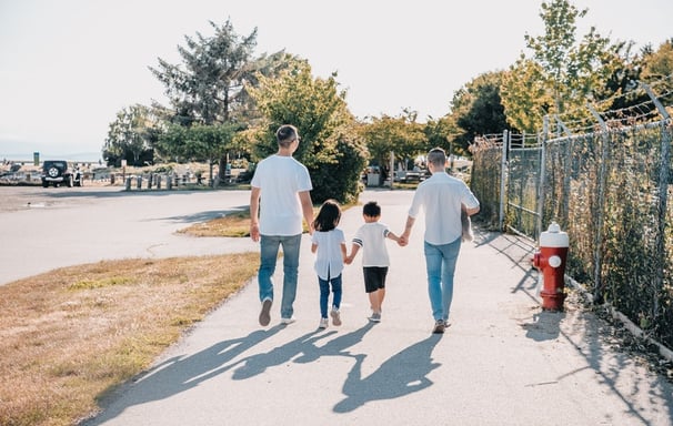 Two dads walking and holding hands with their children on a wide sidewalk in the sun