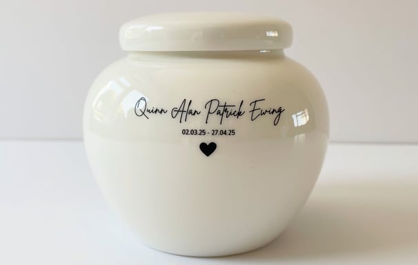 This is a rounded ivory ceramic urn engraved with the text Quinn Alan Patrick Ewing, and adorned with a small heart icon