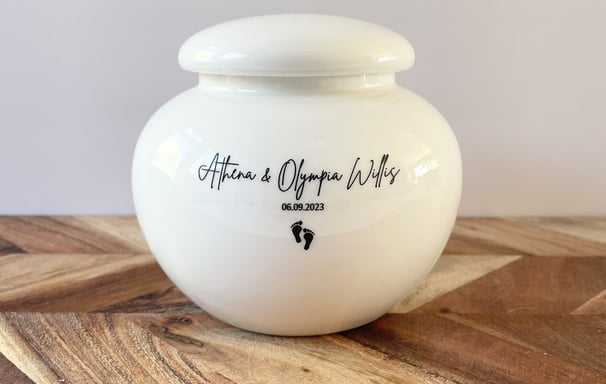 This is a rounded ivory ceramic urn engraved with the text Athena & Olympia Willis, and adorned with a small footprint icon