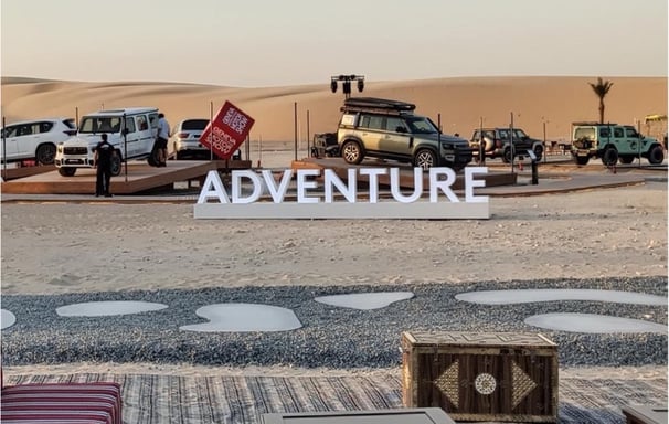 An outdoor car show setup in the desert featuring SUVs, a stage, and a large "ADVENTURE" sign.