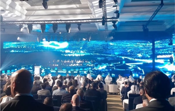 A large conference with an audience, vibrant LED screens, and overhead rigging in a grand venue.