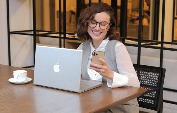 35+ woman mentor smiling, wearing gray, holding phone, sitting with MacBook on table