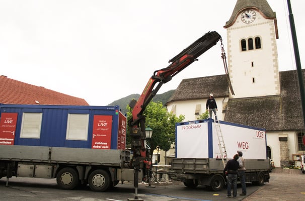 Kran bei der Aufstellung des Ausstellungscontainers "Aus weg los!" auf einem Dorfplatz.