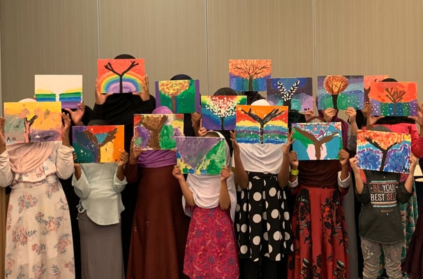 a group of Muslim girls standing in rows, holding colorful tree paintings up, hiding their faces