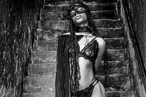 Photo artistique noire et blanc d'une jeune femme noire en sous vêtements et bas qui porte un masque dans le sous sol 