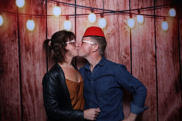 A couple kiss in the get the picture UK vintage photo booth with a wooden backdrop and party glasses