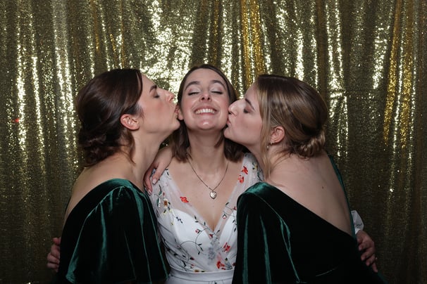 A bride is kissed by two bridesmaids at her wedding event in the get the picture vintage photo booth Brighton golden backdrop