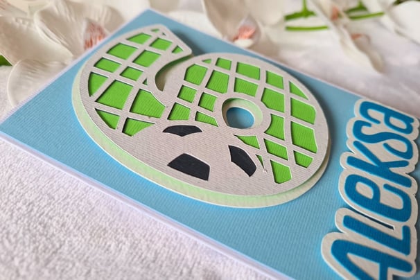 Greeting card with a textured number 6 cut-out of a stylized soccer ball in green and gray. Blue bac