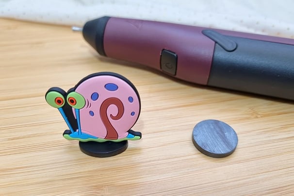 A colorful snail figurine with big eyes next to a hot glue gun and a round magnet on a wooden surfac
