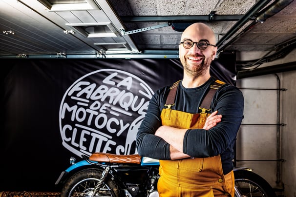 Homme barbu et avec des lunettes debout devant une moto souriant