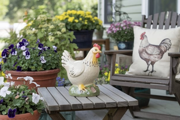 A front porch styled with a  small wooden table holding a ceramic chicken figurine, surrounded by fl