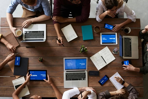 A group of people sitting around a table with laptops, pens, notebook, and cups.