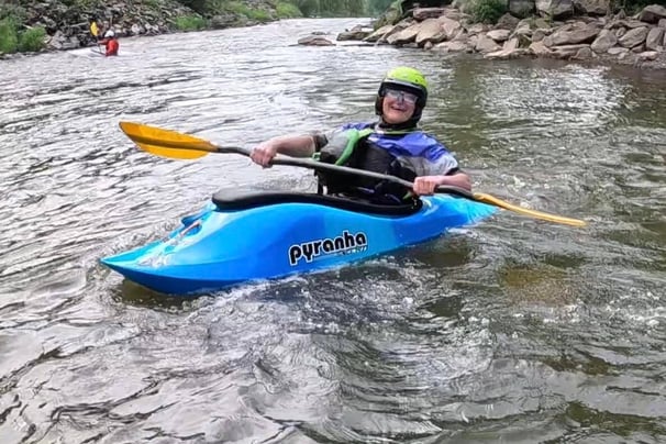 Women in a playboat (freestyle kayak) holding a paddle, Wearing helmet and BA  smiling. 