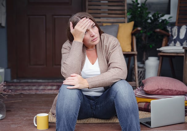 Middle-aged woman sitting on her front porch, hand on forehead, looking tired and reflective. A lapt