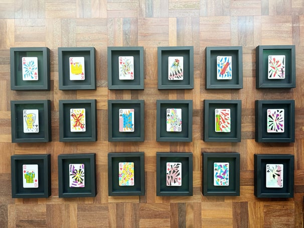 vista de cima das 18 cartas de baralho personalizadas e colecionáveis da série, emolduradas em preto sobre chão de taco.