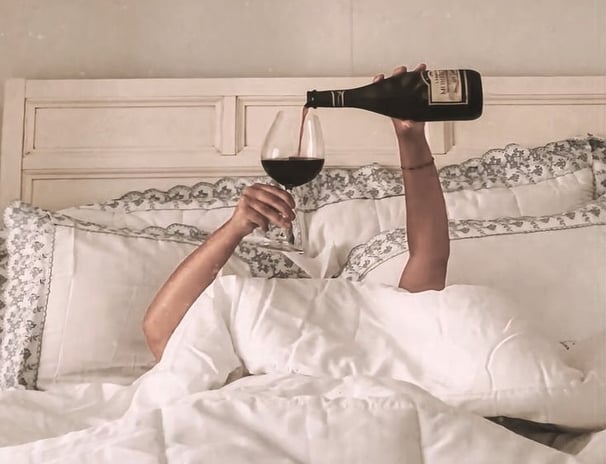 Woman lounging in bed pouring red wine — cozy, indulgent slow living moment