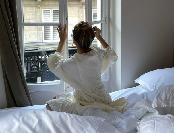 Woman in a robe sitting on a bed and tying her hair in front of a Parisian window — soft morning 