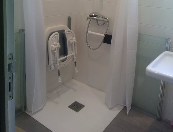Une salle de bains pour personne à mobilité reduite, avec un siège ddouche rabattable, barres de maintient et une paroi à mii