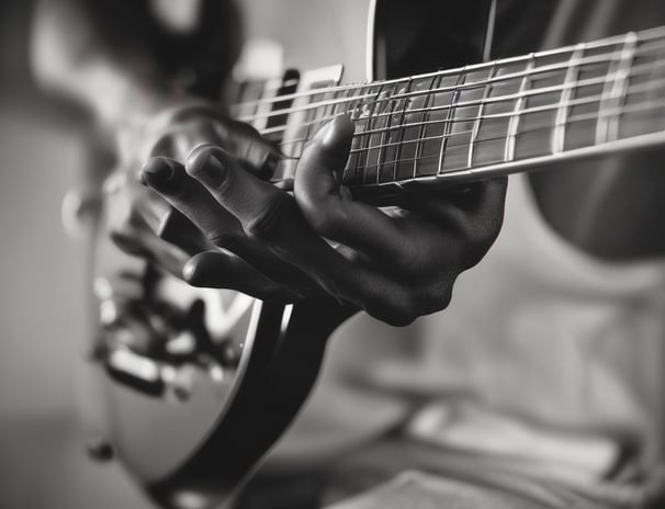 A person is playing an electric guitar. The image is captured in black and white, highlighting the musician's fingers on the fretboard and the guitar headstock with the brand logo visible. The focus is on the hand's positioning and the strings of the guitar.