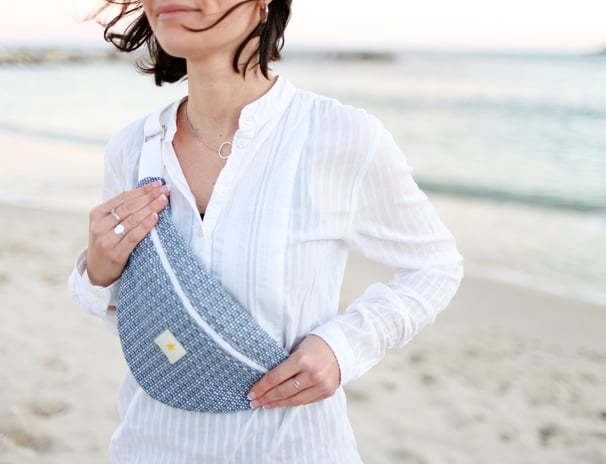 Femme debout à la plage portant un sac banane ÉTOFÉ bleu en tissu revalorisé, tenue blanche estivale