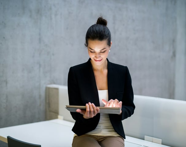 A businesswoman in a black jacket, sitting on a desk, holding a tablet computer.