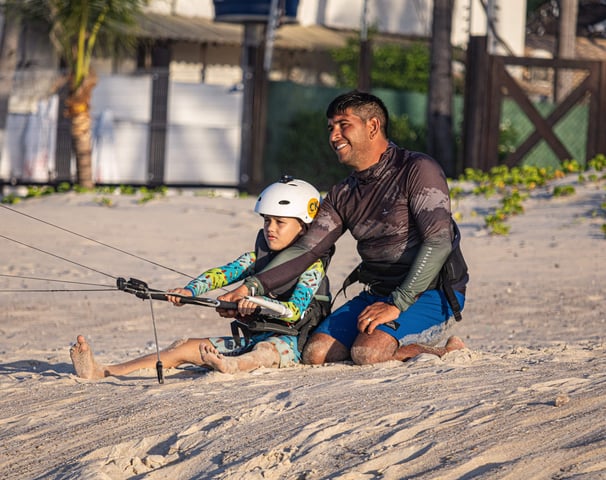 criança sentada na praia aprendendo kitesurf com o instrutor.