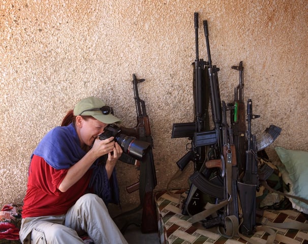 A women holds a camera while sitting next to a pile of guns