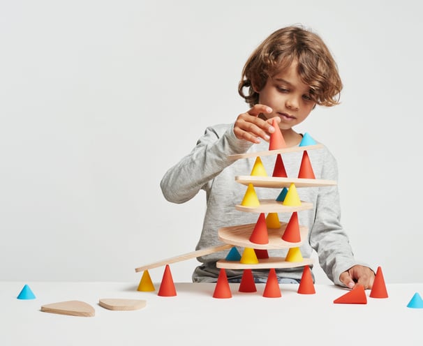 A boy creating a structure from red, blue and yellow silicone cones and wooden tables in the room