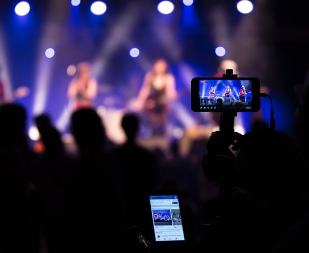 a person holding a cell phone in front of a stage with a video camera live streaming on facebook