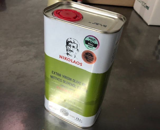 Eine Dose NIKOLAOS Extra Virgin Olive Oil steht auf einer metallischen Oberfläche, mit Preissiegeln 