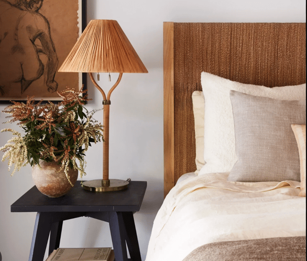 brown lamp on a black table beside cream color bedsheets with a neutral brown bed frame, plant