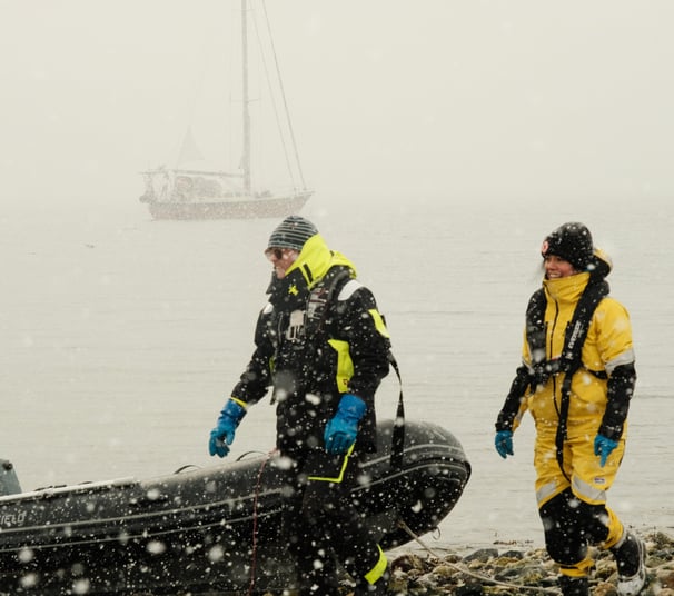 People in a snowy beach with a dinghy and a sailing yacht in the background | Ski and sail in Lyngen