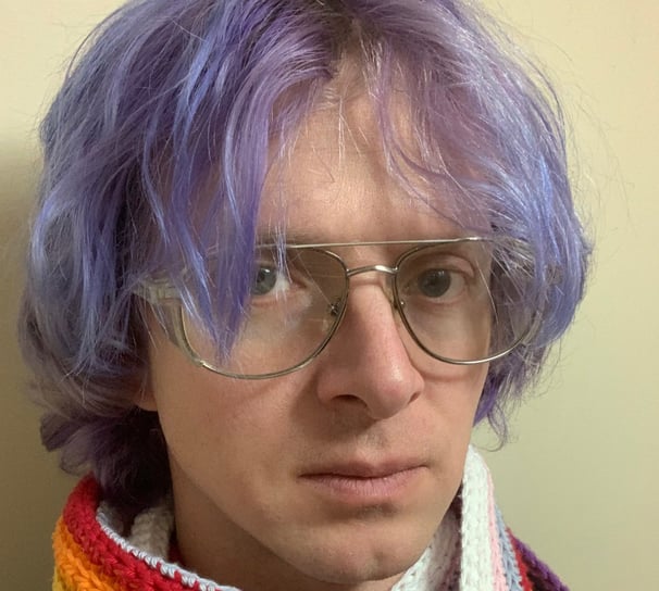 Tomkins with glasses on, wearing a rainbow scarf and with purple hair.