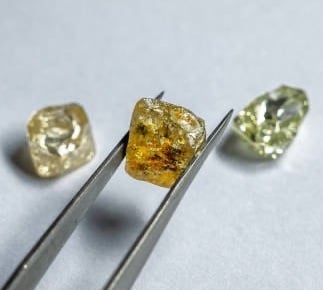 The image features three rough, uncut diamonds resting on a smooth, light-colored surface. A pair of