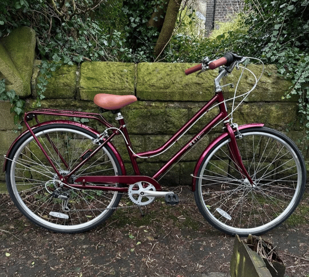 Vélo de ville Reid Classic bordeaux pour femme, avec selle et poignées marron, posé contre un mur de