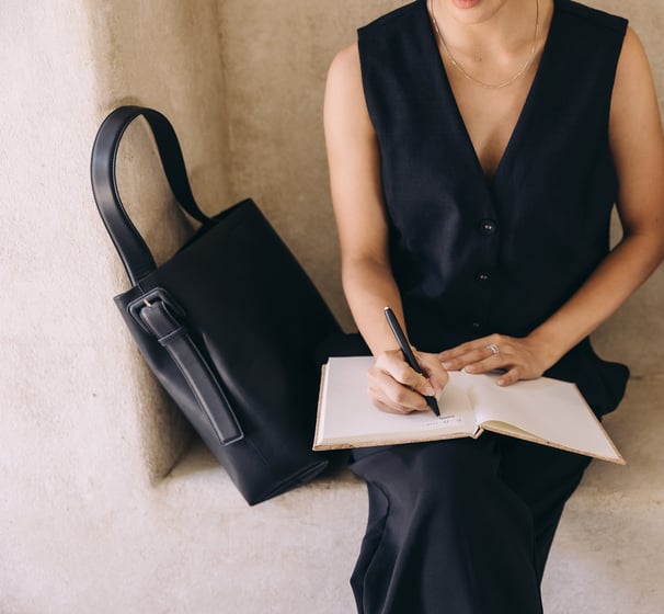 A woman in a sleeveless black dress, sitting and writing in a notebook.