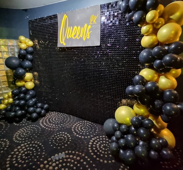 Black shimmer wall with a balloon garland wrapping both sides in black and gold with Queens sign in the middle in Brisbane ev