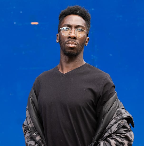 Man wearing circular wireframe glasses, black t-shirt and an Urban camo jacket.